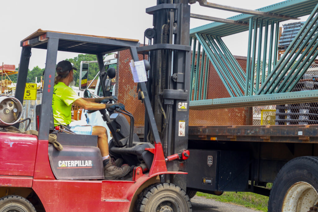 Outside with a worker on a red and black forklift loading racking materials onto a flatbed truck