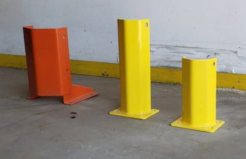 Angled photo of rack protectors in various sizes, yellow and orange, on the floor of a warehouse near a white wall.