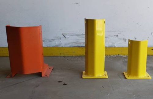 Rack protectors in various sizes, yellow and orange, on the floor of a warehouse near a white wall.