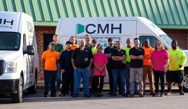 A photo of the Coastal Material Handling staff in front of two work vans with the Coastal Material Handling logo. The photo is taken in front of the Coastal Material Handling offices in Suffolk, Virginia.