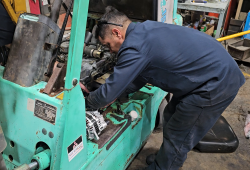 A forklift mechanic working on a green forklift. The seat has been removed and he is working on the engine of the forklift.